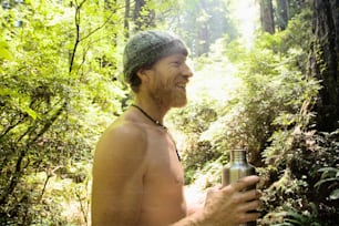 Ein Mann ohne Hemd, der ein Bier in einem Wald hält