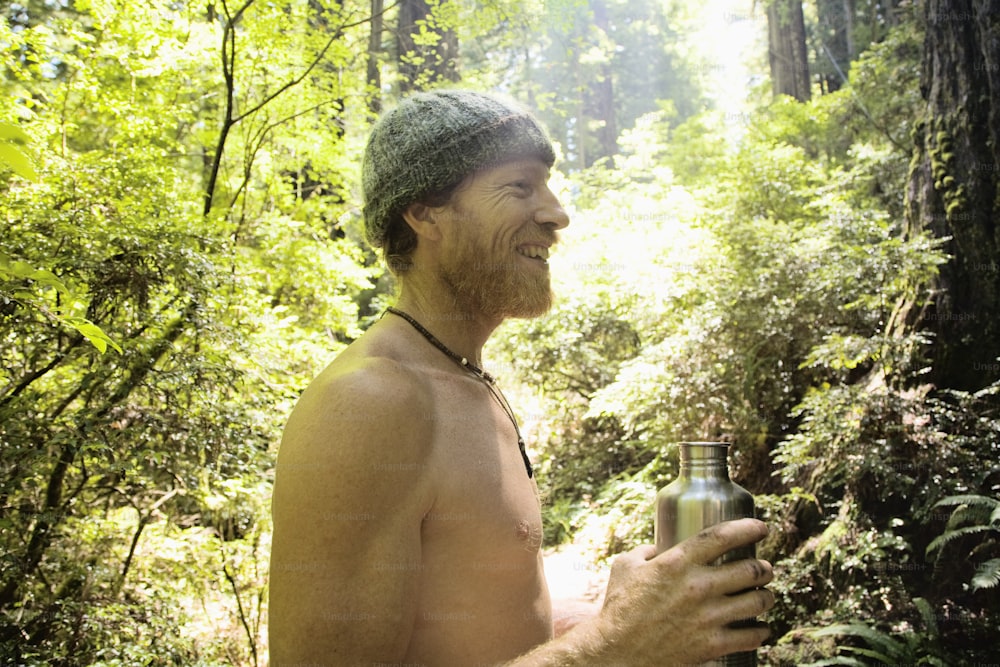Ein Mann ohne Hemd, der ein Bier in einem Wald hält