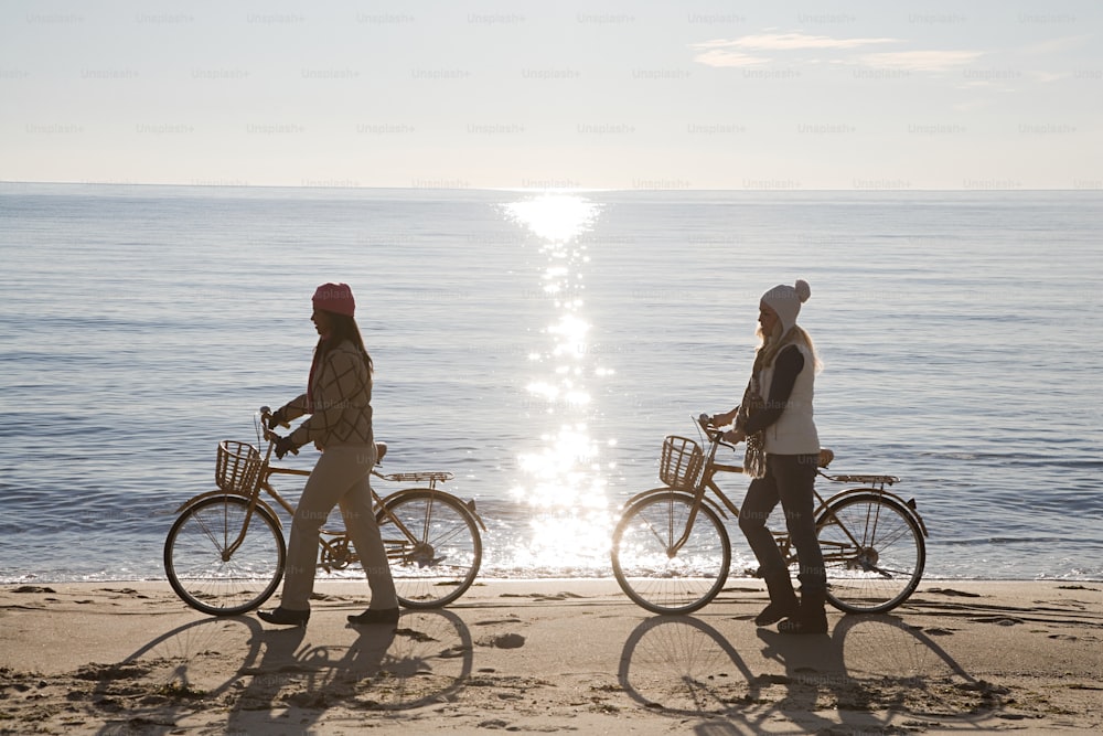 two people walking on the beach with their bikes