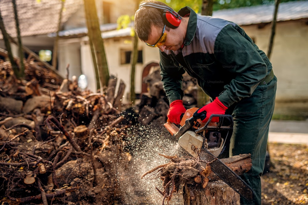 Foto ravvicinata di un boscaiolo in uniforme e protezione che lavora su un mucchio di legna con una motosega nel cortile della casa in una giornata di sole.