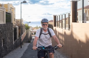 スポーツヘルメットとサングラスをかけて、夕暮れ時に上り坂の路地で電動自転車をサイクリングするシニアの笑顔の男性。背景に水面の地平線