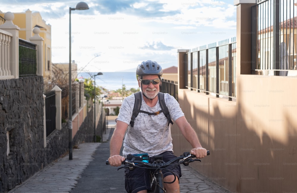 Homem sorridente sênior com capacete esportivo e óculos de sol pedalando com sua bicicleta elétrica no beco de subida ao pôr do sol. Horizonte sobre a água no fundo