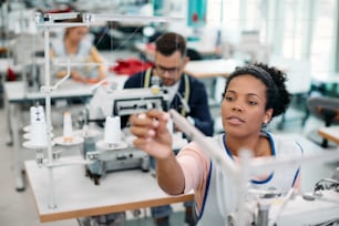 Afroamerikanische Näherin setzt den Faden auf die Nähmaschine, während sie an der Produktionslinie in der Textilfabrik arbeitet.