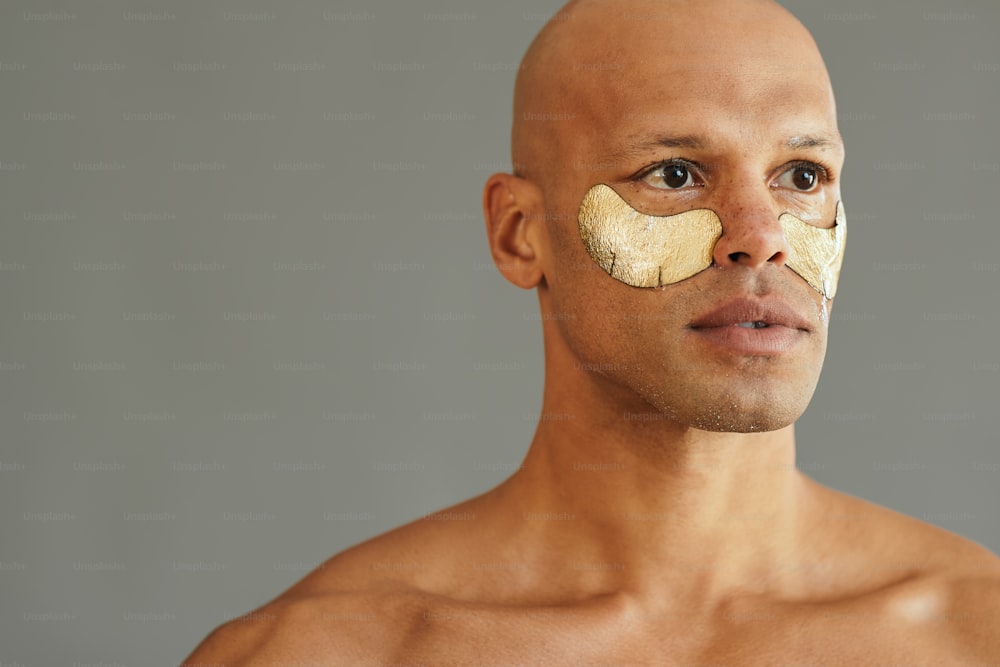 Young African American man using golden under-eye recovery patches during facial skin treatment. Copy space.