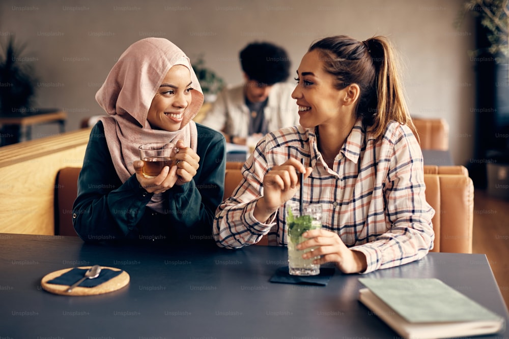 Young happy Muslim woman and her Caucasian female friend communicating while sitting in a cafe.