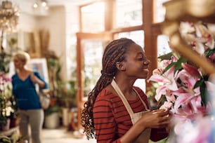 Happy African American florist smelling fresh flowers while working at flower shop.