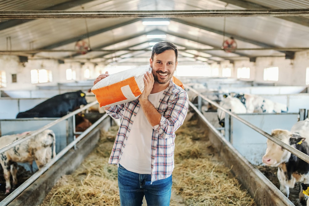 Joven granjero caucásico atractivo sonriente de pie en el granero con un saco con comida para animales sobre el hombro y mirando a la cámara.