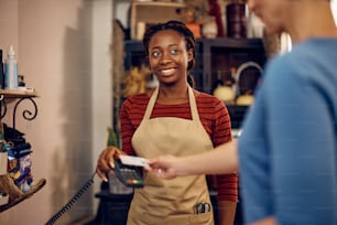 Feliz florista afroamericana sosteniendo un lector de tarjetas de crédito mientras su cliente paga sin contacto en una floristería.