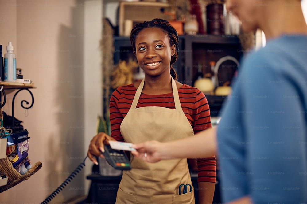 Happy African American florist holding credit card reader while her customer is paying contactless at flower shop.