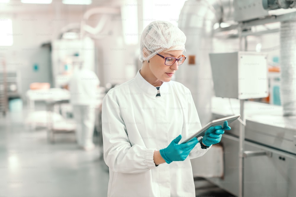 Supervisor in sterile uniform and with eyeglasses using tablet for controlling workflow in food factory.