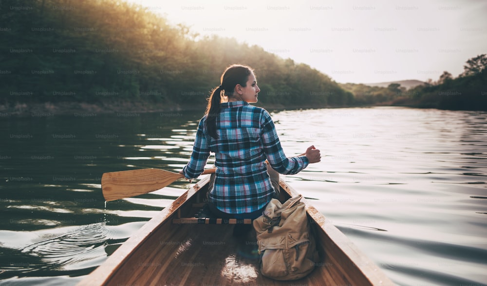 Rear view of hipster girl paddling the canoe on the sunset lake.