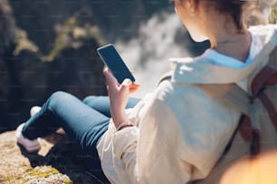 Beautiful traveling girl sitting on top of mountain at sunset and using mobile phone.