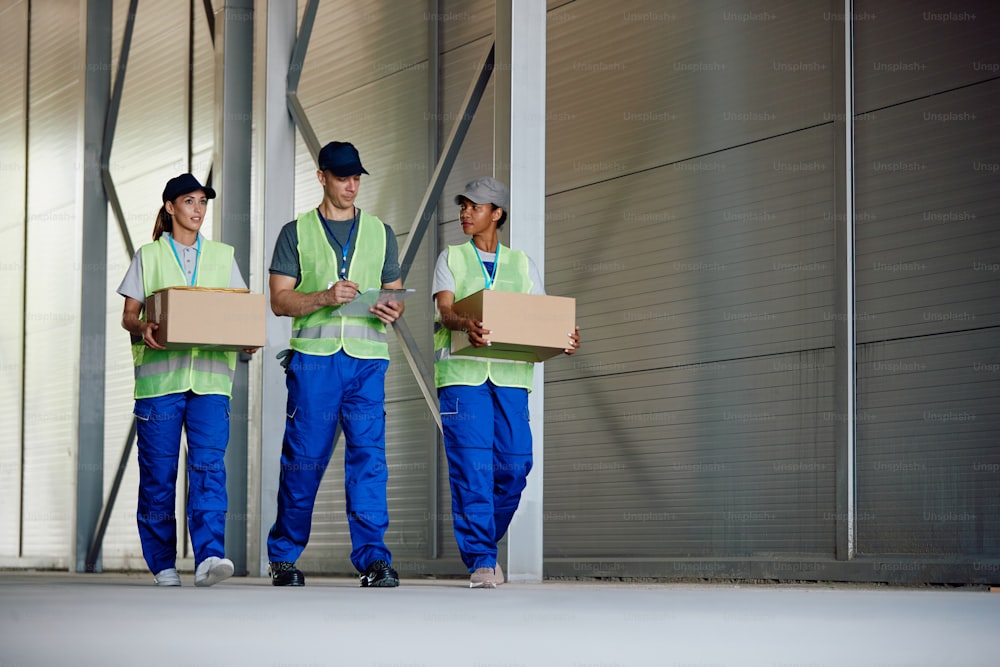 Warehouse manager analyzing check list while female workers are carrying cardboard boxes at storage compartment. Copy space.