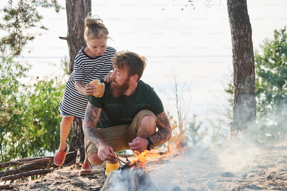 Happy family basking near the fire at the cozy forest. Charming girl looking with love at her father while giving sandwich to him. Stock photo