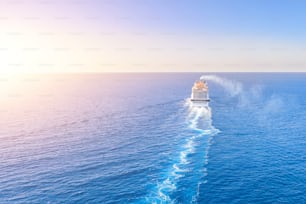 Le paquebot de croisière va à l’horizon de la mer bleue laissant un panache à la surface du paysage marin au coucher du soleil. Vue aérienne, concept de voyage en mer, croisières