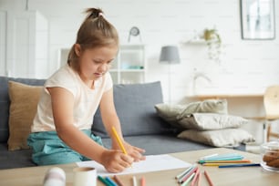 Menina com síndrome de down sentada à mesa e desenho da imagem com lápis coloridos na sala de estar em casa