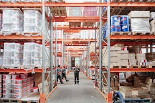Young intercultural warehouse workers in uniform moving along aisle between large racks while male manager with tablet standing on crossing