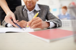 Primer plano de un niño africano sentado en el escritorio y trabajando en su cuaderno con el profesor señalando el error durante una lección
