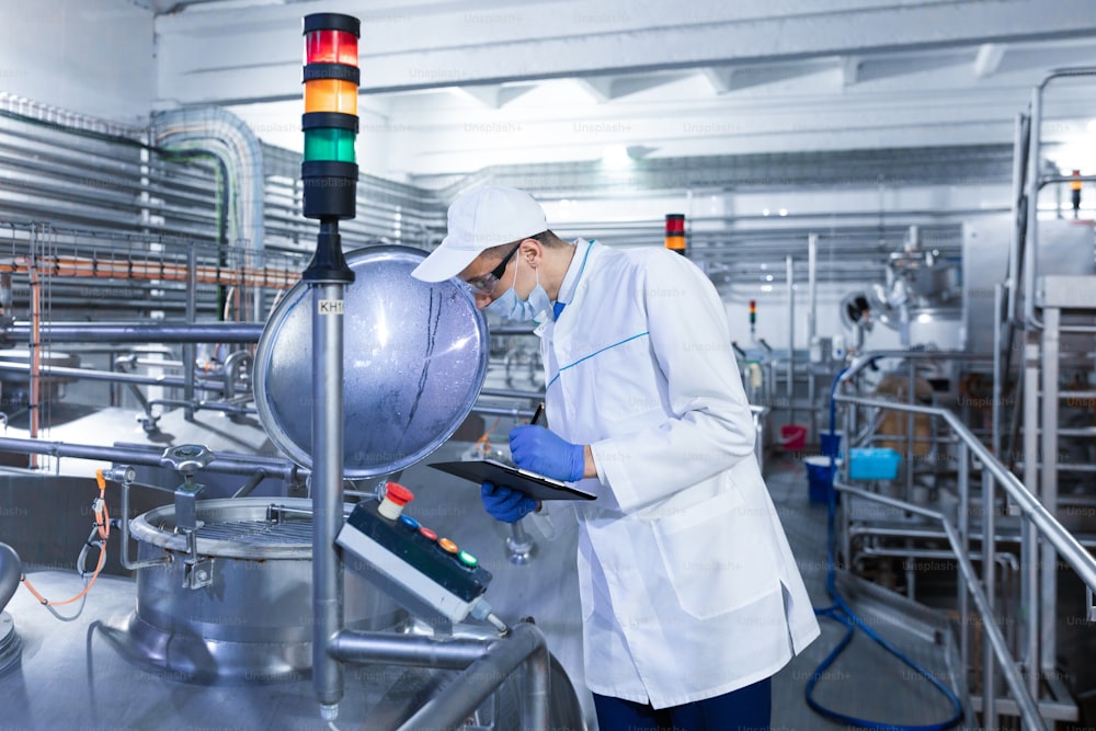 technologist in the cap, mask and white coat stands in the production shop and makes notes with a pen in the sheet. The inspector removes the indicators at the dairy plant. Engineer keeps statistics on production