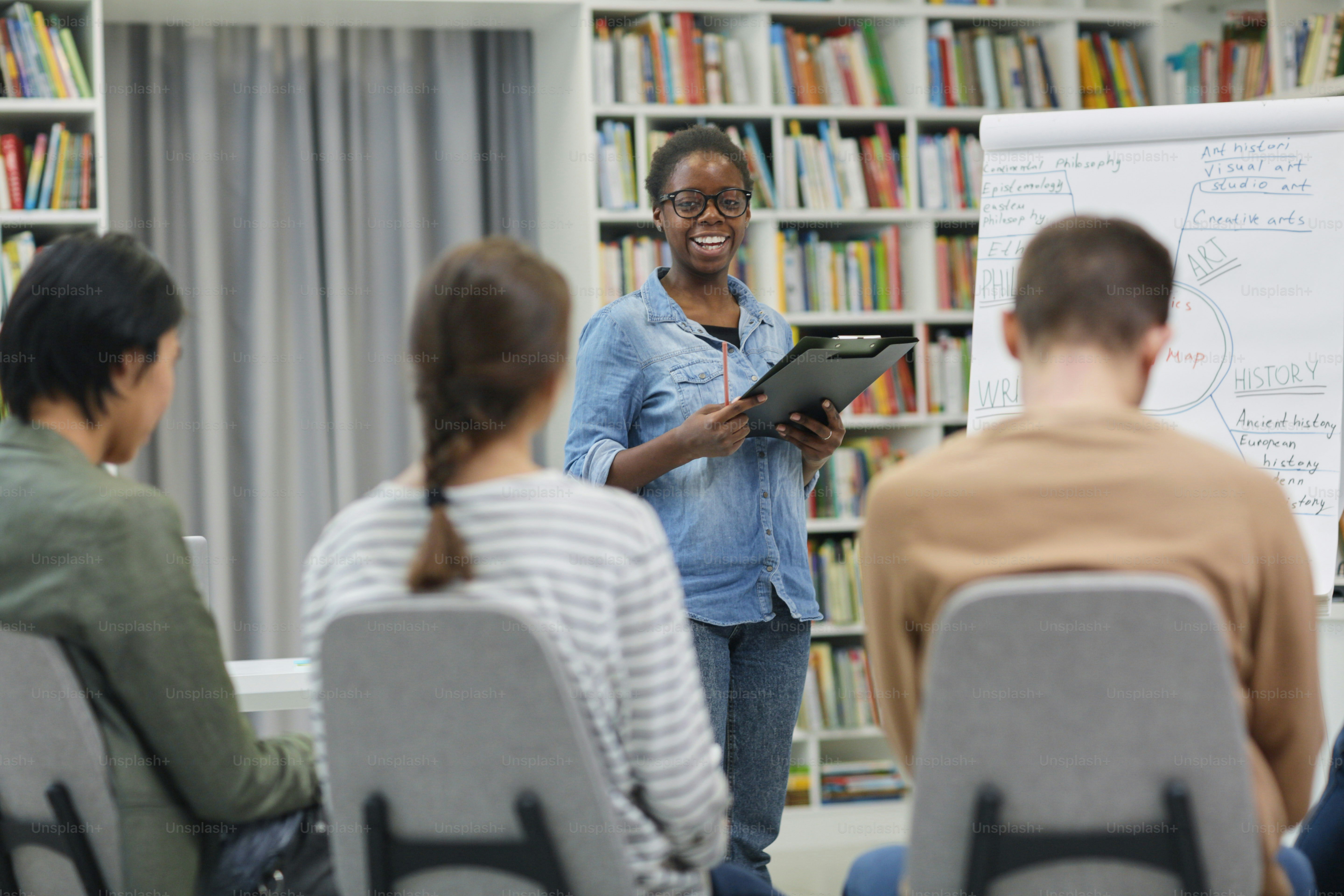 African smiling woman standing near the whiteboard and presenting her presentation to young people in the library