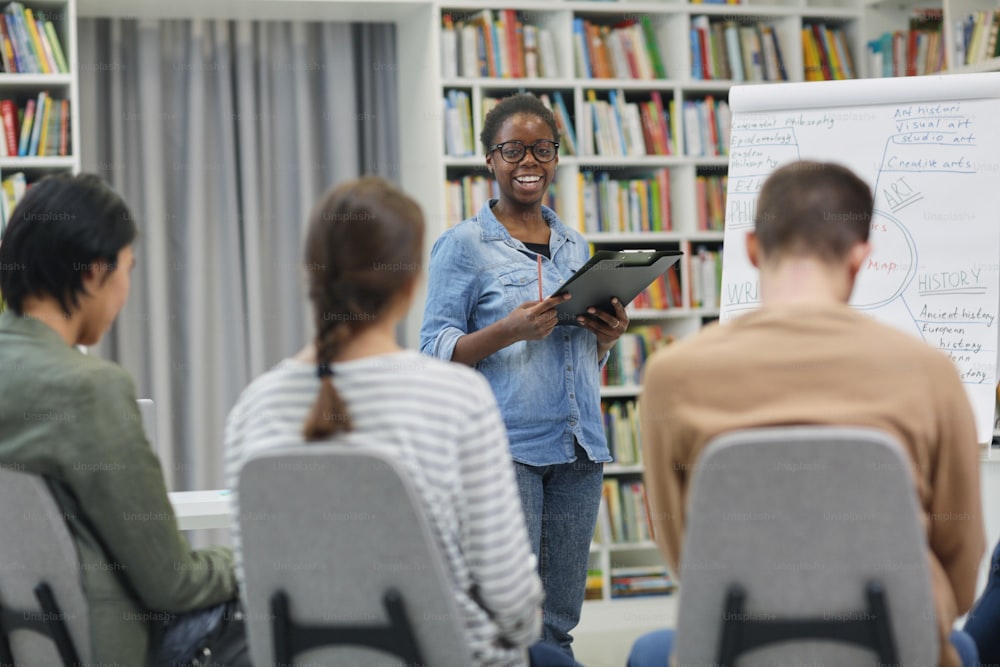 Mujer africana sonriente de pie cerca de la pizarra y presentando su presentación a los jóvenes en la biblioteca