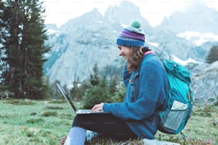 Young beautiful traveling woman with backpack, hat and laptop sitting on rock and trying to connect network in the stunning mountain wilderness in front of amazing cold lake.