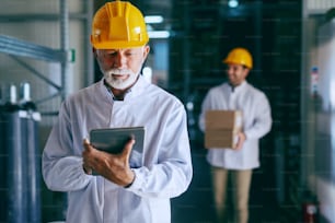 Retrato de un trabajador de almacén adulto mayor con uniforme blanco y casco en la cabeza usando tableta. En el fondo, un trabajador más joven que lleva cajas con mercancías.