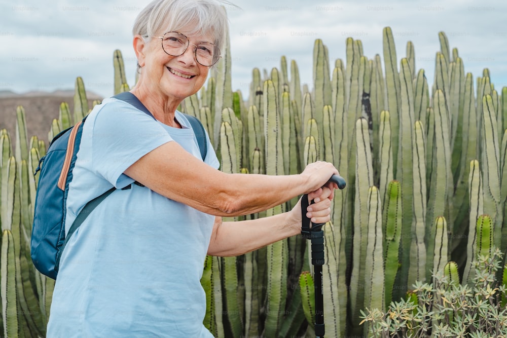 Donna anziana allegra durante un'escursione all'aperto che trasporta uno zaino e un bastone da passeggio mentre riposa vicino a un grande cactus. Un'anziana donna in forma durante un'escursione che si gode la libertà dell'avventura e una vacanza salutare