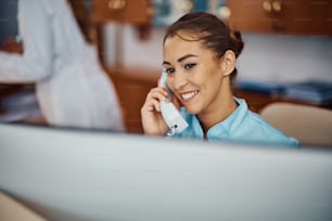 Young nurse answering a phone call while working on desktop PC at reception desk in a hospital. Copy space.