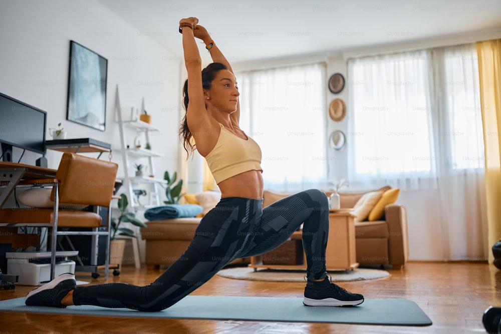 Female athlete in deep lunge pose holding arms above her head during home workout.