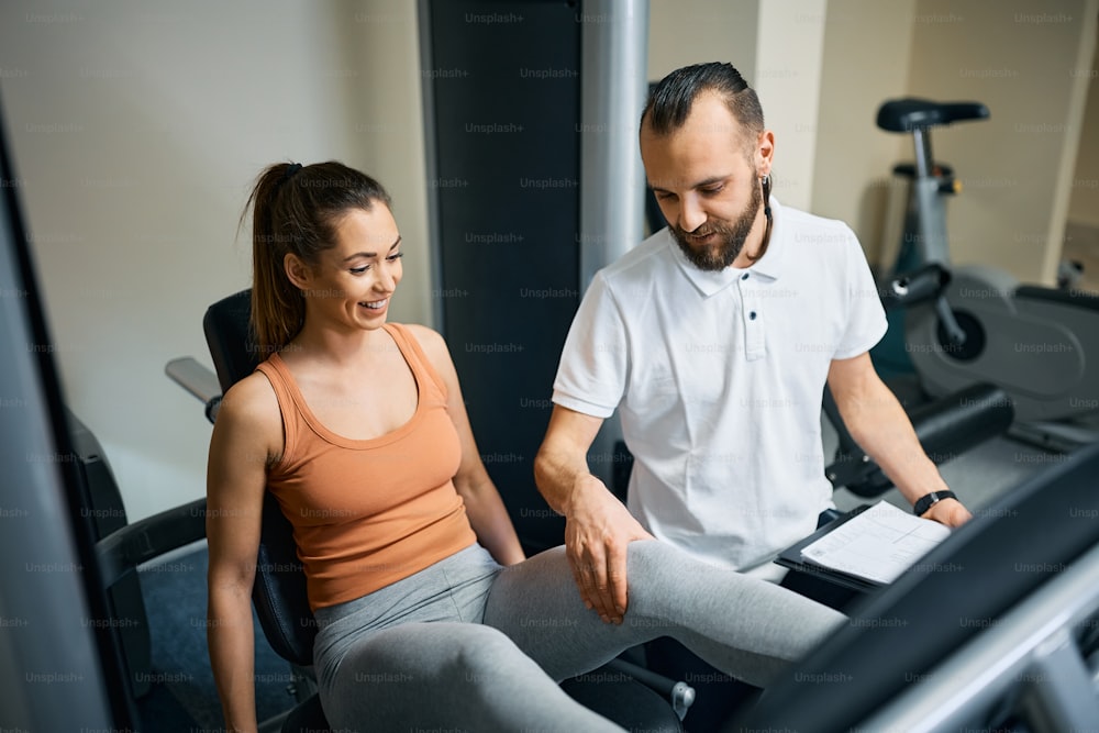 Happy personal trainer assisting athletic woman with exercising on leg press machine in health club.