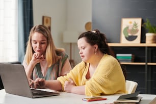 Pensive Caucasian woman and her co-worker with Down syndrome sitting in front of laptop working on business project