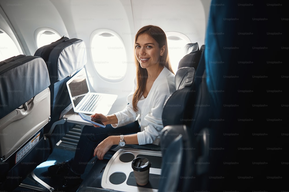 Pasajera segura de sí misma sentada junto al ojo de buey del avión con una computadora portátil frente a ella y un teléfono inteligente en las manos