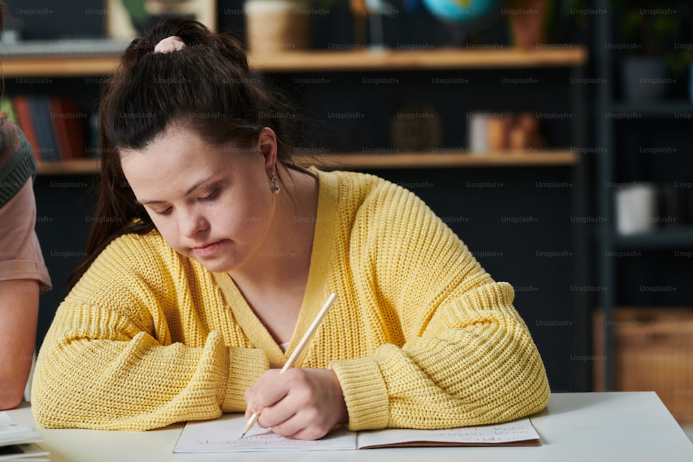 Caucasian girl with Down syndrome sitting at table writing something in workbook during individual class at home