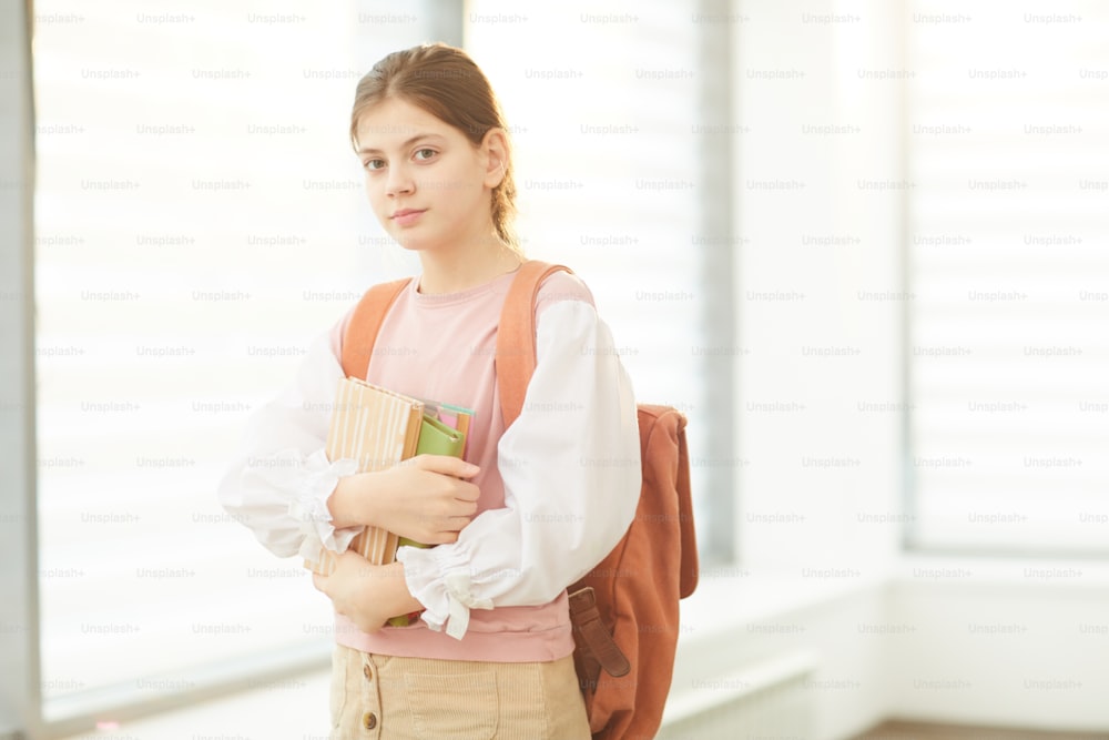 Horizontal medium portrait of beautiful Caucasian teen girl wearing backpack holding books looking at camera, copy space