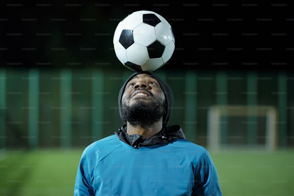 Bearded blackman in sports uniform looking at soccer ball on his forehead while training at stadium before match