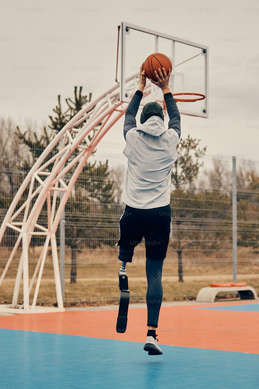 Back view athletic man with artificial leg shooting at the hoop while playing basketball on outdoor court.