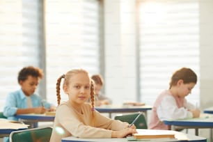 Horizontal portrait shot of little Caucasian girl with blond hair sitting at school desk holding pencil looking at camera, copy space