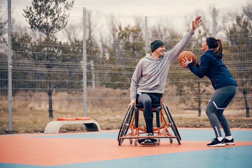 屋外のスポーツコートで車椅子の友人とバスケットボールをしながらシュートを打つ運動女性。