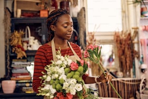 Mulher afro-americana usando flores frescas enquanto faz buquê no trabalho na floricultura.
