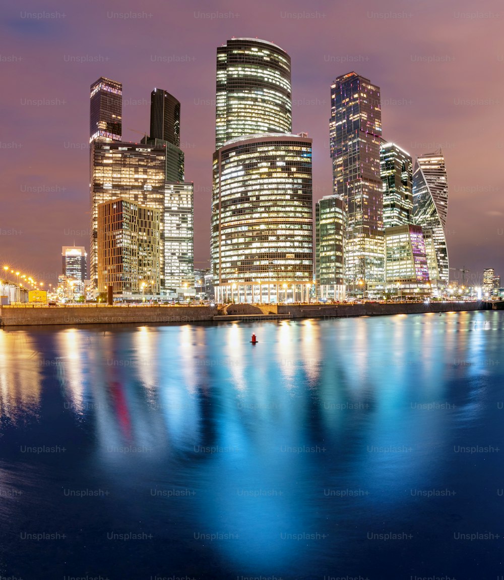 Beleuchtete Wolkenkratzer in Moskau City oder internationales Geschäftszentrum bei Nacht mit Lichtern, Blick vom Wasserteichdamm mit Reflexionen