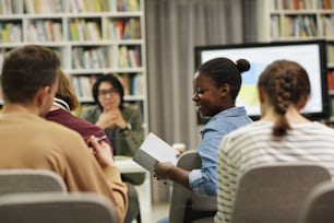 African young woman smiling and reading a book and discussing it with her colleagues during presentation in the library
