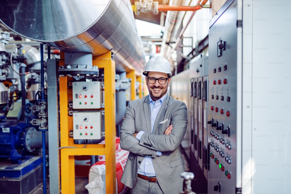 Smiling handsome caucasian businessman in gray suit and with helmet on head standing with arms folded next to dashboard. Power plant interior.