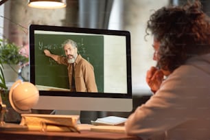 Rear view of young man looking at computer monitor and studying online while teacher telling something and pointing at blackboard