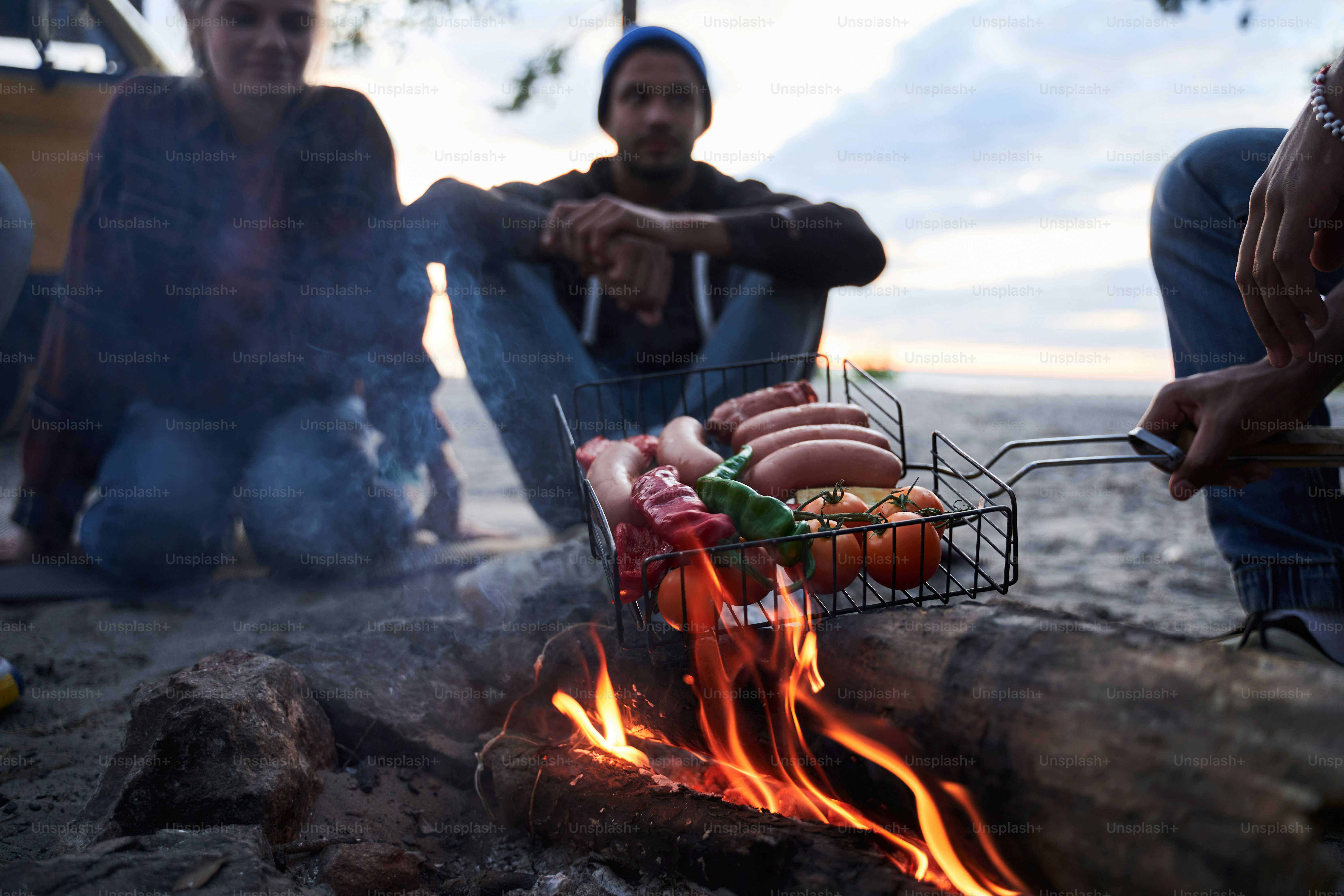 Friends having fun a barbecue party in nature. People sitting in a circle around the fire while waiting for dinner