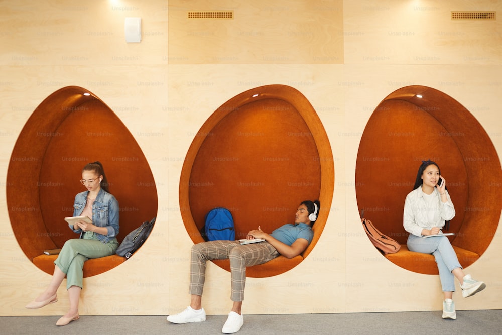 Modern multi-ethnic students in casual outfits sitting in egg-shaped wall chairs and using gadgets during break