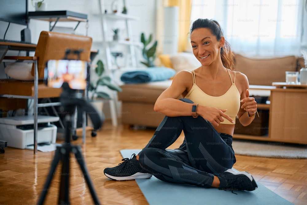 Happy female fitness instructor talking while doing stretching exercises and live streaming from home.