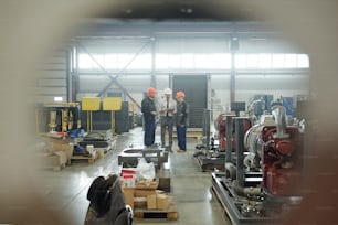 Three engineers of contemporary industrial plant standing in the center of workshop and discussing technical sketch in touchpad