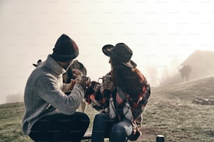 Young couple holding cups and looking at cow while sitting on the valley in mountains