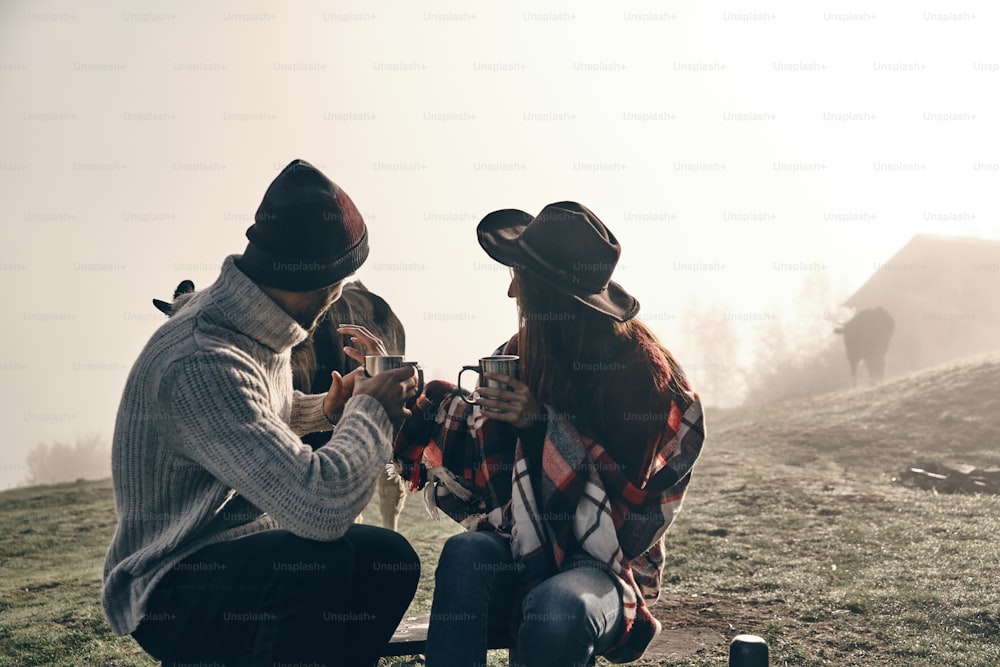 Young couple holding cups and looking at cow while sitting on the valley in mountains